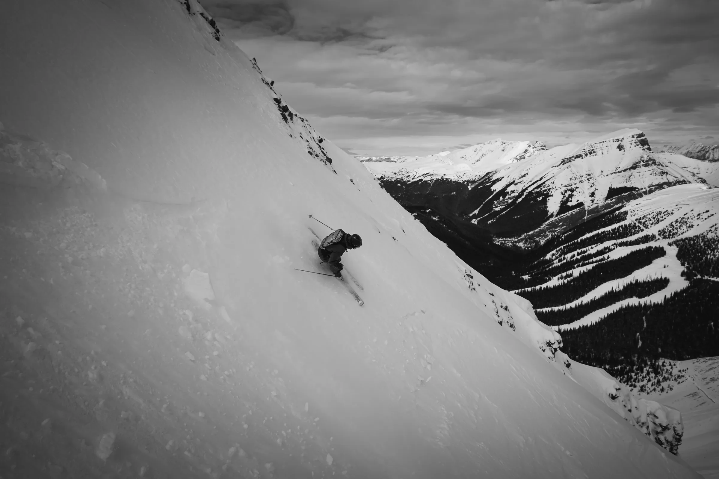A skier crossing beneath a broad mountain face in black and white