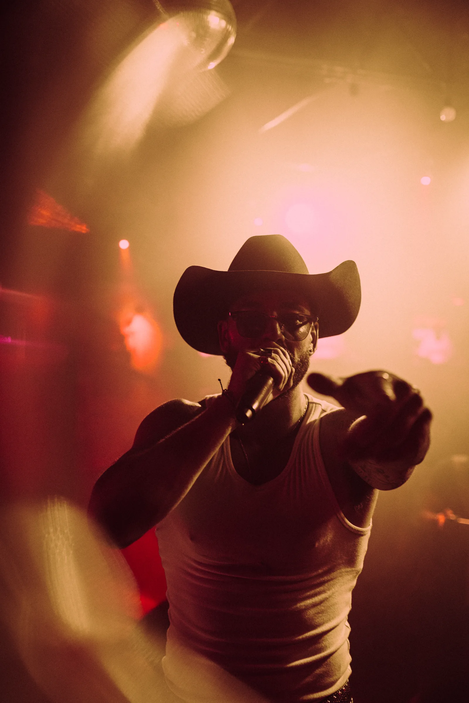 A performer in a cowboy hat pointing toward the camera under amber light
